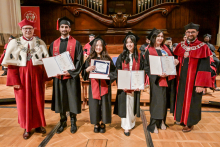 Six people in togas and birettas stand on stage. Four people in the centre hold diplomas. At the sides people in red togas with white elements and rector's chains. In the background a wooden wall with an organ. Wooden floor.