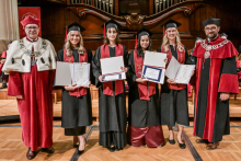 Five people in togas and birettas stand on the wooden floor. Three people in the middle have black togas with red sashes and are holding diplomas. To the sides, people in red togas with white elements and chains. In the background is a wooden organ and a red plaque with the university's coat of arms.