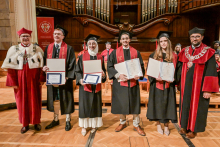 Six people stand on the stage in the auditorium. The four people in the centre have black togas and birettas, holding diplomas. To the sides, people in red togas with rector's insignia. In the background a large wooden organ and seated participants in the ceremony.