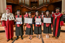 Four graduates in black togas and birettas stand on stage, holding diplomas. To the sides, two men in red togas with rector's insignia. In the background is a wooden organ prospectus.