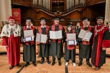 Six graduates in black togas and birettas hold their diplomas. They stand against the backdrop of the wooden organ in the concert hall. To their sides, two men in red togas with rector's insignia.