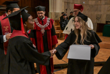 A female student wearing a black toga and biretta with a red accent shakes hands with the person presenting the diploma. The person is wearing a black toga and holding a white document. In the background, the ceremony participants can be seen wearing togas with red elements and decorative chains.