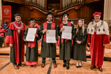 Six people in togas and birettas stand on stage. Three people in the middle hold diplomas. On the left, a person in a red toga with a chain. On the right, a person in a red toga with a fur collar. In the background a large wooden organ.