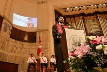 An alumnus in a black toga and biretta stands at the lectern with the logo “Universitas Medica Anon”. In the background, a screen with the words “Congratulations to Muhammad Mostafa Metwally”. On the left, a flag and three men in ceremonial dress. On the right, a bouquet of flowers.