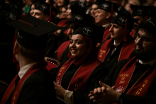 A group of graduates in black togas and birettas sit during the graduation ceremony. They have maroon sashes with the words ‘CLASS OF 2025’ written on them. Their faces are blurred. Other participants in the event are visible in the background.