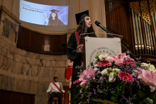 In the foreground, a podium with a microphone and a bouquet of flowers. Behind the podium stands a person in a black toga and biretta, speaking. In the background, a screen with the words “Agendas 6th Year Class Representative Alexandra Pissa” and a photo of a person in a biretta. To the left sits a person in historical costume.
