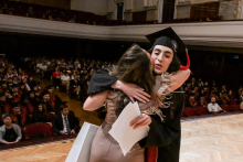 Two women embrace on stage during a graduation ceremony. One of them is wearing a black toga and red biretta, holding a diploma. The audience seated in the large hall can be seen in the background.