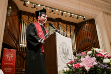 An alumnus in a black toga and biretta speaks on a podium with the logo of the Warsaw Medical University. The organ and golden-brown curtains are visible in the background. To the left stands a red flag with the university's coat of arms. In the foreground are flowers in pink, white and purple.