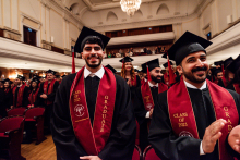 A group of graduates in black togas and birettas stand in the concert hall. They have red sashes with the words “CLASS OF 2025” and “GRADUATE” written on them. The balcony with the audience is visible in the background.