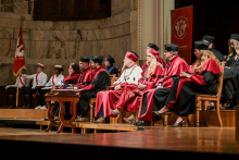 A group of people in red and black togas and birettas sit on stage during an academic ceremony. In the background a marble wall and a banner with the university crest. To the left, people dressed in white with red sashes.