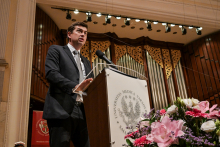 A man in a suit stands at the lectern with the iWUM coat of arms. The organ and decorations are visible in the background. To the right a bouquet of flowers in pink and white.