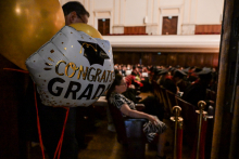 The auditorium during the graduation ceremony. In the foreground is a white balloon with the words “CONGRATS GRAD” and a black graduation cap. In the background people are seated, some in togas and birettas. The walls are covered with wooden panels, the ceiling bright and high.