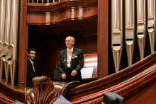 Two men in elegant suits stand on a wooden balcony next to silver organ pipes. An older man in a black dinner jacket, white shirt and bow tie stands in the foreground. The other man is partially obscured. In the background is an open sheet music book on a lectern. The dominant colours are brown and silver.