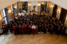 A large group of graduates in black togas and birettas stand in an elegant room with wooden floors and chandeliers. They hold their diplomas and raise their birettas. In the background are mirrors and people watching the ceremony. In the foreground people in red togas.
