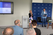 In the foreground, people sitting with their backs. In the middle of the room, a woman, the pro-rector, with a microphone and a man with documents stand next to a lectern with a logo. The woman is dressed in a colourful dress and black jumper. The man is wearing a blue jacket, white shirt and dark trousers. In the background is a screen and a blue wall with a repeating logo and text.