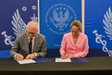 A man in a grey suit and a woman in a pink jacket are signing documents at a table.
