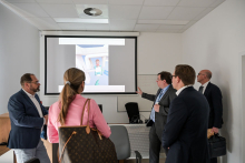 Five people are in a conference room – three standing, two sitting – watching a presentation on a projection screen.