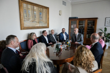 Several people are seated at an oval table in a formal meeting room with drinks on the table and books in the background.