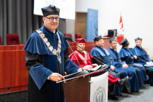 Graduation ceremony for nursing and midwifery graduates. In the foreground, a person wearing a gown and cap is standing at a lectern with a microphone, giving a speech. In the background, several other people, also dressed in gowns, are sitting.