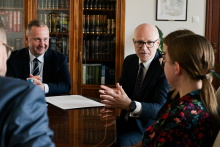 Four people are sitting at a table in a conference room. One of them is addressing the others, gesturing.