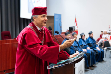 Graduation ceremony for nursing and midwifery graduates. In the foreground, a man in a red gown and biretta is speaking into a microphone. The man is wearing a rector's chain. In the background, several people in blue gowns are seated.