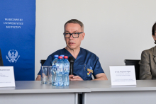 A man in medical attire is sitting at a table. In front of him are bottles of water and glasses. On the left, there is a blue banner with the inscription “Medical University of Warsaw” and an emblem. On the table are two nameplates, one with the inscription “Dr. Wojciech Figiel.”