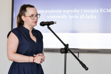 A woman in a navy blue dress stands at the microphone, holding her hands folded in front of her.