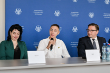 Three people are sitting at a table with name tags reading “Wiktoria” and “Danił.” The person in the middle is holding a microphone. There are bottles of water on the table. In the background, the logo and the words “Warsaw Medical University” are repeated multiple times.