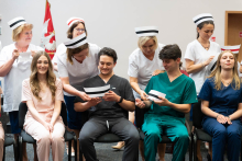 Graduation ceremony for nursing and midwifery students. Four people in colorful medical uniforms (two young women and two young men) are sitting in the front row. Behind them are four people in white uniforms who are placing nursing caps on the heads of those seated.