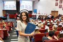 Graduation ceremony for nursing and midwifery graduates. A young woman in a beige medical uniform and nursing cap stands in the middle of the auditorium, holding a blue folder with the words “Medical University of Warsaw” written on it.