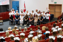 Graduation ceremony for nursing and midwifery students. A group of young women in colorful medical uniforms and nursing caps sit on the stage in the auditorium. Behind them stand older women in white uniforms, placing nursing caps on the heads of those seated.
