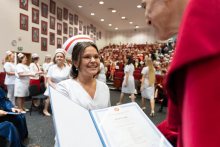Graduation ceremony for nursing and midwifery graduates. A man in a red gown presents a diploma to a young woman in a white medical uniform and midwife's cap.