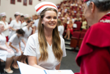 Graduation ceremony for nursing and midwifery graduates. A woman in a white medical uniform and midwife's cap receives a document from an elderly man in a red academic gown.