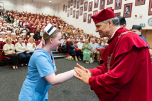 Graduation ceremony for nursing and midwifery graduates. A woman in a blue medical uniform and midwife's cap stands on stage and shakes hands with an elderly man in a red academic gown.