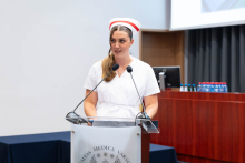 Graduation ceremony for nursing and midwifery graduates. A woman in a white medical uniform and midwife's cap stands behind a lectern with two microphones.