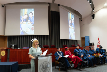 Graduation ceremony for nursing and midwifery graduates. A woman in a green medical uniform and a nurse's cap stands behind a lectern with two microphones, holding sheets of paper.