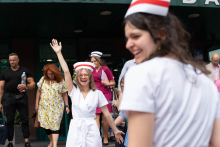 A woman in a white medical uniform and midwife's cap stands outside the building with her hand raised. In the foreground, another woman in a white medical uniform and midwife's cap looks in her direction.