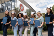 Seven women in medical uniforms and nurse caps stand in front of a building with a large logo of the Medical University of Warsaw. Each of them is holding a navy blue briefcase.
