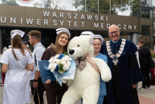 Four people are standing in front of the Medical University of Warsaw building. A woman in a medical uniform and nurse's cap is holding a navy blue briefcase and a bouquet of flowers. Next to her stands a woman holding a large teddy bear and a man in a navy blue academic gown.