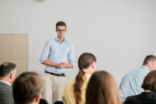 A man wearing glasses speaks to an audience seated in chairs.