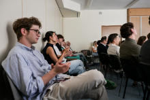 A group of people are sitting in a conference room, facing a speaker who is off-camera. They are dressed casually. Some of them are applauding. In the background, there are light-colored walls and doors.