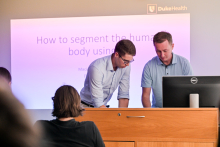 A man wearing glasses and a man giving a lecture are talking at a lectern. In the background, there is a presentation on the screen.