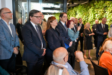 Grupa osób w formalnych strojach stoi i siedzi w eleganckim wnętrzu. W tle widoczna zielona ściana z roślinami.