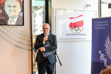 Mężczyzna w średnim wieku, rektor prof. Rafał Krenke, stoi przy mikrofonie. Jest ubrany formalnie. W tle widoczny portret starszego mężczyzny, kolorowe przewody na ścianie, tablica z logo PKOl oraz baner WUM.