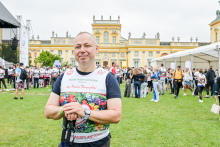 Mężczyzna w koszulce z napisem „POLSKA TRANSPLANTOLOGIA” i kolorowym wzorem stoi na trawie przed żółtym pałacem z zielonymi okiennicami. W tle widać grupę osób w sportowych strojach. Po lewej stronie znajduje się scena z czarnym dachem, po prawej biały namiot. Niebo jest pochmurne.