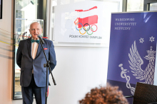 Starszy mężczyzna, prof. Krawczyk, ubrany formalnie przemawia do mikrofonu. W tle po lewej plakat z logo PKOl z konturem Polski i kołami olimpijskimi. Po prawej niebieski baner Warszawskiego Uniwersytetu Medycznego z białym orłem i laską Eskulapa. Zdjęcie wykonane w pomieszczeniu przy oknie.