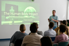 A man in a blue shirt presents “The Human Body Project.” A white skeleton and a medical device are visible on the screen. There are seven people in the room. The room is bright, with white walls.
