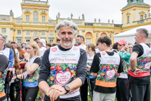 Około dwudziestu osób bierze udział w biegu charytatywnym na tle żółtego pałacu. Mężczyzna z siwymi włosami na pierwszym planie sprawdza zegarek. Uczestnicy noszą kolorowe koszulki z napisem „Bieg po nowe życie”. W tle kobiety i mężczyźni przygotowują się do startu.