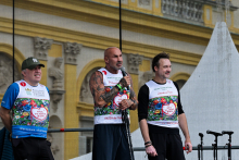 Trzech mężczyzn stoi na scenie przed budynkiem z żółtymi i białymi elementami. Noszą białe koszulki z kolorowym nadrukiem i napisem „Bieg po nowe życie”. Mężczyzna po lewej ma niebieską koszulkę i zieloną czapkę. Środkowy trzyma mikrofon, ma tatuaże i biało-czerwoną opaskę. Mężczyzna po prawej nosi czarną koszulkę z długim rękawem.