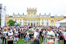 About forty people are standing on the grass in front of a yellow and white palace building. The women and men are wearing colorful sports shirts and holding Nordic walking poles. On the left side, there is a white banner with logos, and on the right side, there is a white tent.